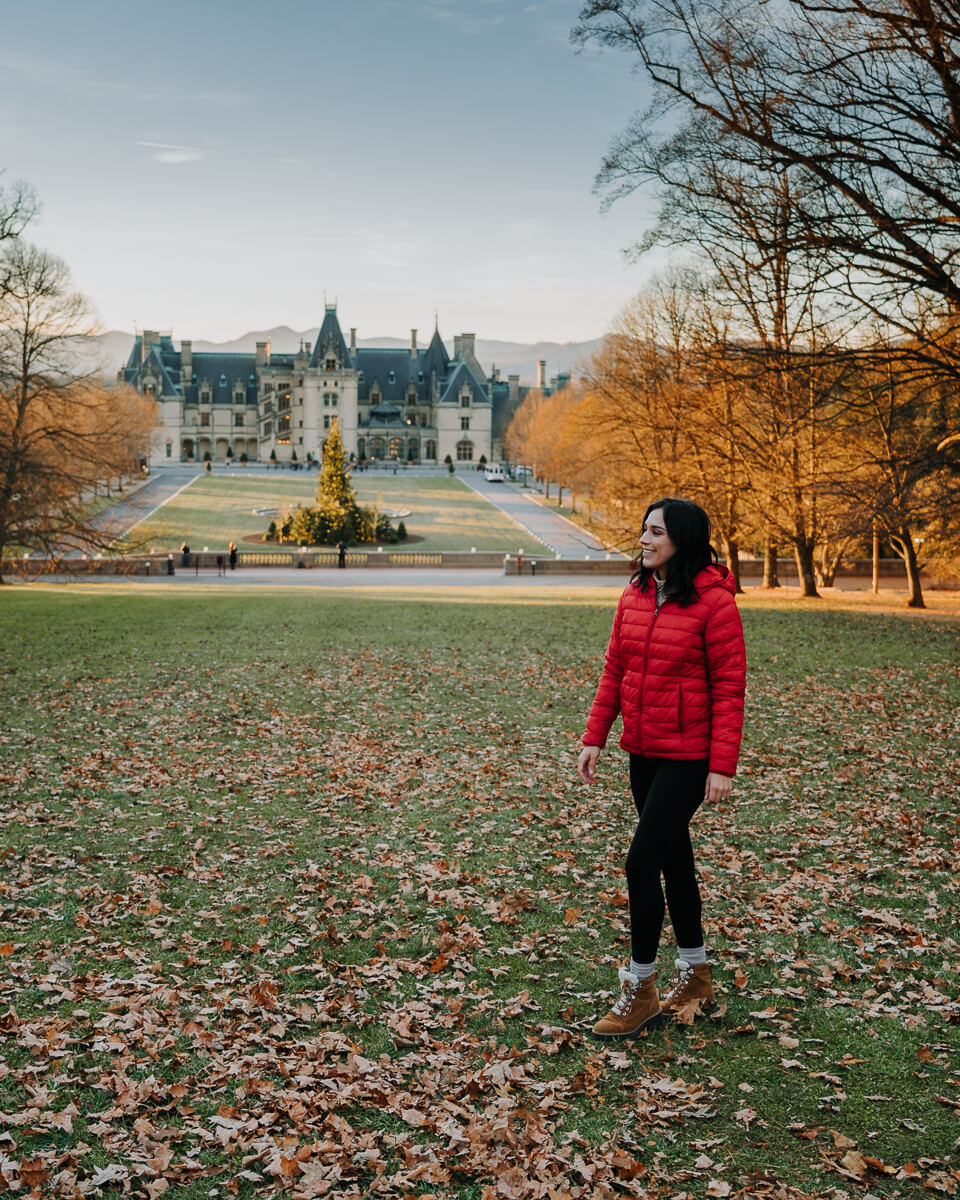 Woman in red jacket stands in a field with Biltmore House and the mountains off in the distance