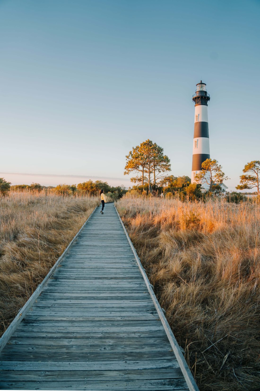 Woman standing on boardwalk in front of Bodie Island Lighthouse on a sunny morning